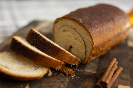 A sliced loaf of cinnamon swirl bread on a wooden cutting board with cinnamon sticks beside it.