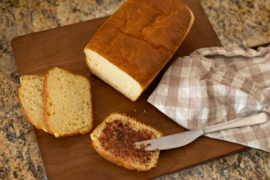 A loaf of bread on a wooden board, with two slices cut. One slice is spread with a brown topping. A plaid cloth and butter knife are nearby.