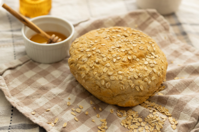 Round oat bread on a checkered cloth next to a small dish of honey with a honey dipper.