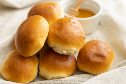 A stack of golden brown bread rolls on a cloth, with a small white bowl of honey in the background.