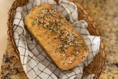 Loaf of multigrain bread topped with seeds in a wicker basket lined with a checkered cloth on a granite surface.