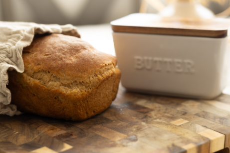 Loaf of bread partially covered with cloth next to a butter container on a wooden surface.