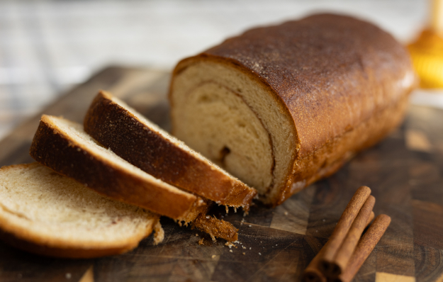 A loaf of cinnamon swirl bread with several slices cut on a wooden cutting board, accompanied by two cinnamon sticks.