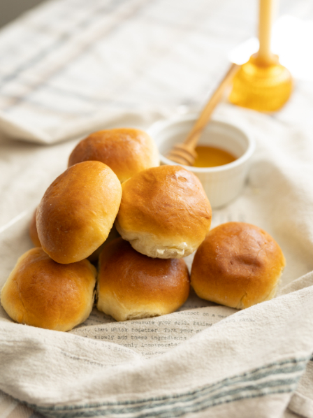 A stack of six golden brown dinner rolls on a cloth, with a small bowl of honey and a honey dipper in the background.