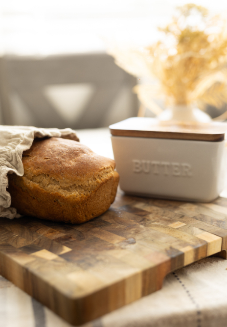 Loaf of bread partially covered with cloth on a wooden cutting board. Butter dish and vase with dried flowers in the background.